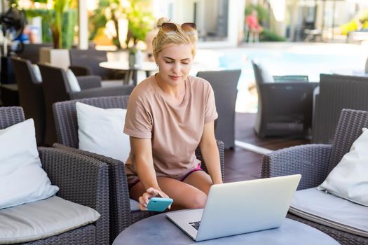 Young female sitting working on laptop in public wifi area, typing, people passing by on the background.