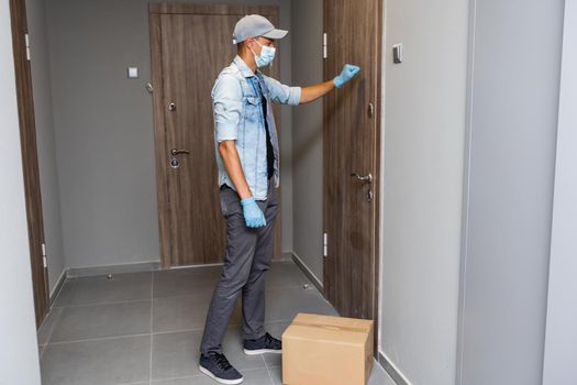 Delivery. Cheerful young courier holding a cardboard box while standing at the entrance of apartment.