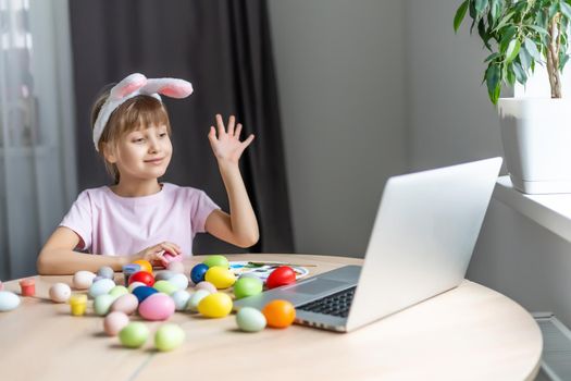 Pretty little girl wearing bunny ears and painting easter eggs at home during coronavirus covid-19 outbreak. Kid using laptop on kitchen, online, video call to friends.