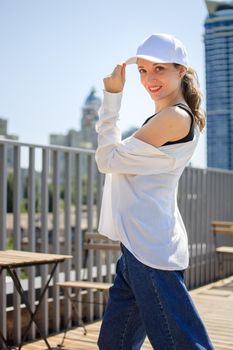 Female portrait of young active girl in black top, white shirt, basketball cap, and jeans on modern buildings background outdoors.