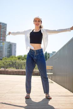 Female portrait of young active girl in black top, white shirt, basketball cap, and jeans on modern buildings background outdoors.
