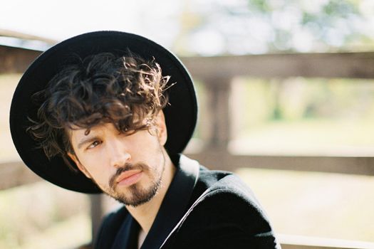 Young man in a black hat sits near a wooden railing. Portrait. High quality photo