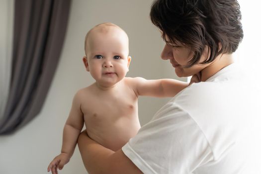 Mother and child on a white bed. Mom and baby boy in diaper playing in bedroom. Parent and little kid relaxing at home. Family having fun together