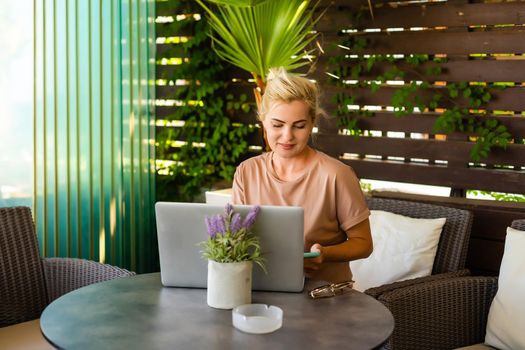 A side view photo of woman using laptop for e-shopping while sitting in a modern hotel lobby. A blonde woman wearing dress is looking at the screen.