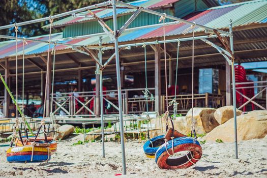Hang rope swing old tires paint bright multicolor orange blue yellow red metal frame veranda roof. Nature sand beach background. Day sun summer. Child kindergarten joy sandbox playground.
