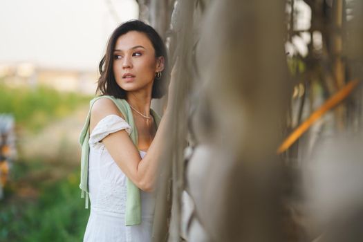 Pretty young Asian woman, posing near a tobacco drying shed, wearing a white dress and green wellies. Beauty and fashion concept