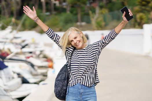 Woman in her 60s strolling along a seaside spot near the beach, raising her arms in joy. Elderly female enjoying her free time.