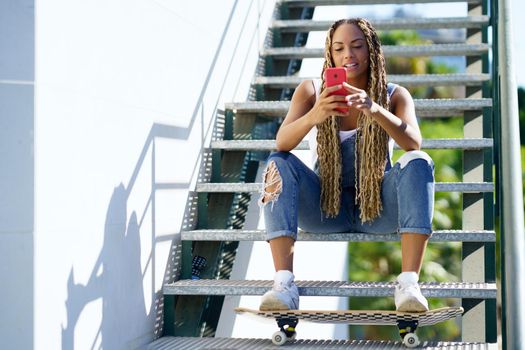 Young black woman with coloured braids, sitting on some steps while consulting her smartphone with her feet resting on a skateboard.