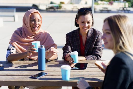 Happy young multiethnic female friends, in casual clothes, smiling while resting in outdoor cafe and drinking coffee from take-away cups
