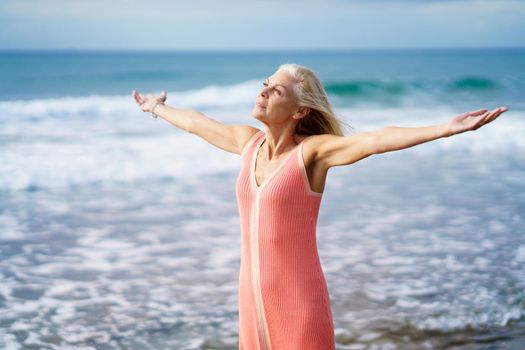 Beautiful mature woman opening her arms on a beautiful beach, spending her leisure time. Elderly female enjoying her retirement at a seaside retreat.