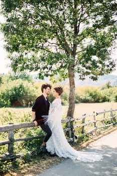 Bride stands near groom sitting on a wooden fence near a country road. High quality photo