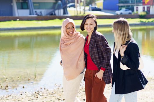 Cheerful young multiracial female friends in stylish clothes smiling and chatting while walking together on embankment near river on sunny day in city