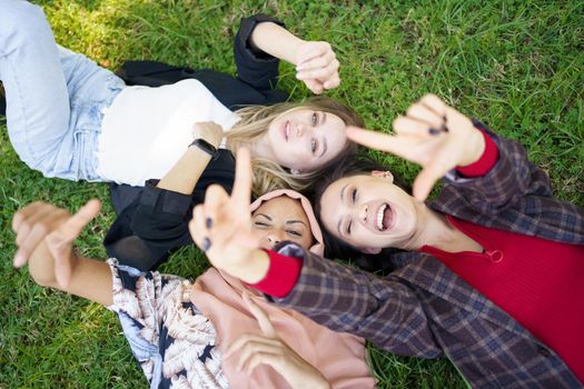 Top view of playful young diverse female friends in casual clothes laughing while lying on grassy lawn and making camera gesture with hands