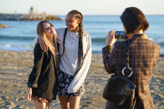 Female taking picture on cellphone of smiling multiethnic women hugging each other while standing on sandy seashore at sunset