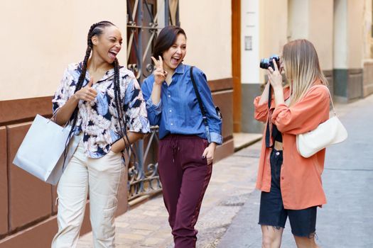 Joyful young multiracial female tourists, smiling and winking while showing v sign and thumb up gesture, during walk on city street with female friend taking picture on photo camera