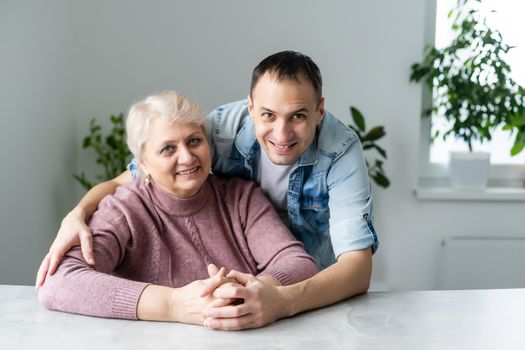 Head shot happy young bearded man embracing beautiful smiling middle aged senior mother in eyewear, relaxing together on comfortable sofa in living room, enjoying funny conversation or gossiping.