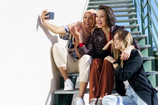 Trendy young cheerful Muslim lady in stylish outfit and traditional hijab, smiling brightly while taking selfie on mobile phone, sitting on staircase with diverse girlfriends on sunny day