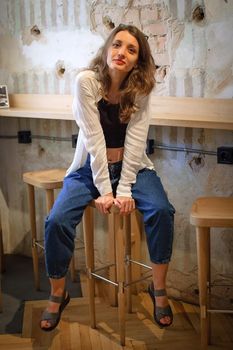 Indoor female portrait of a playful girl in black top, white shirt, and jeans sitting on wall background in loft style cafe.