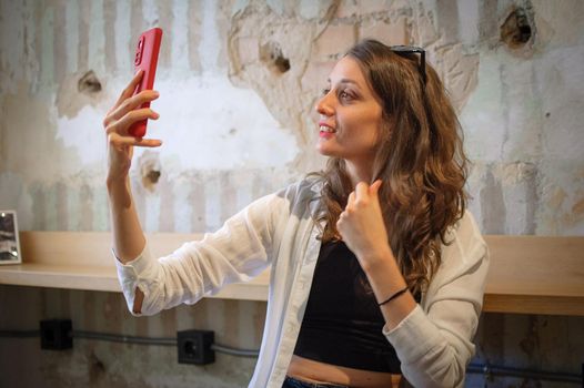 Female portrait of young active girl in white shirt taking a photo of herself indoors on loft wall background, selfie time, happy people concept