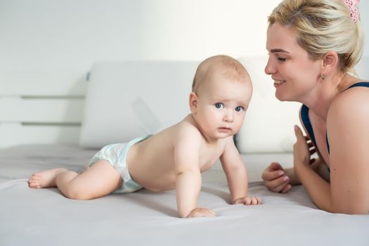 Mother and child on a white bed. Mom and baby boy in diaper playing in sunny bedroom. Parent and little kid relaxing at home. Family having fun together. Bedding and textile for infant nursery.