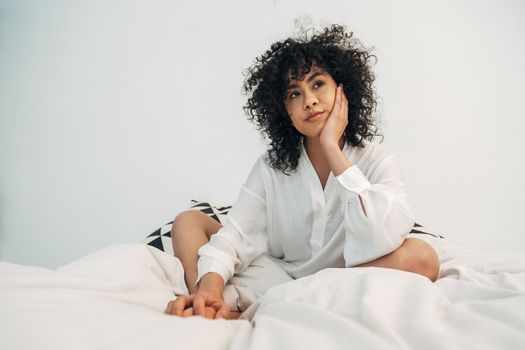 Pensive young mixed race woman with curly hair sitting on bed resting face on hand. Copy space. Lifestyle concept.