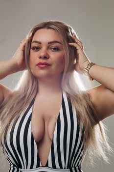 Gorgeous blonde lady wearing a striped one-piece swimsuit and holding hands behind her head while posing for camera