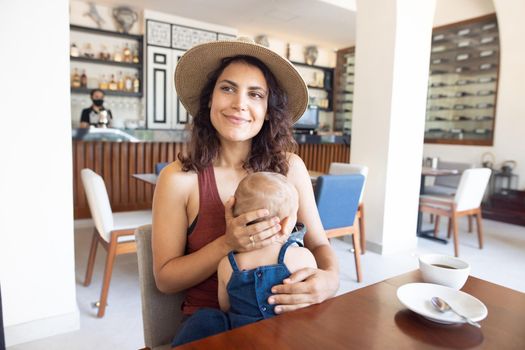 Portrait of beautiful smiling mother wearing a hat and holding adorable baby in restaurant. Happy brunette woman and young daughter at table with cafe counter as background. Lovely family on holiday