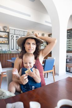 Portrait of beautiful holding her hat and cute happy baby in restaurant. Attractive brunette woman and adorable young daughter at table with cafe counter as background. Lovely family on holiday