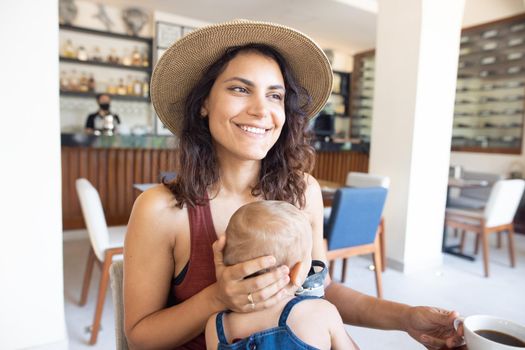 Portrait of beautiful smiling mother with hat holding cute baby and coffee mug in restaurant. Happy brunette woman and young daughter at table with cafe counter as background. Family having breakfast