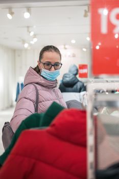 Woman wearing protective face mask shopping clothes in retail shopping store. Concept of lifestyle during corona virus pandemic.