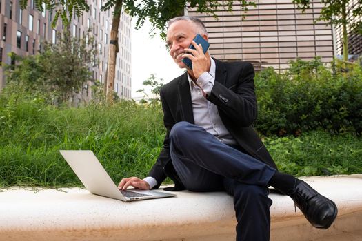 Caucasian businessman using laptop and talking on mobile phone sitting on park bench next to office buildings. Business and technology concept.