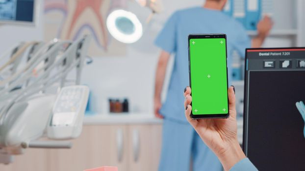 Close up of dentist holding smartphone with green screen for teethcare at dental cabinet. Dentistry specialist looking at chroma key and isolated mockup template on mobile phone.