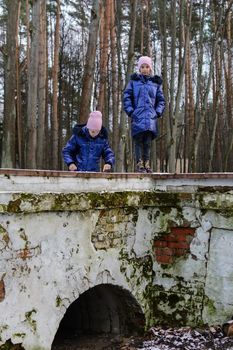 Two twin caucasian girls in blue coats playing on the old bridge in late autumn in the forest