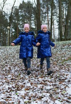 The first frost on autumn. Two happy twin girls in blue coats hand in hand runing down the hill covered with the dry leaves and first snow in the forest