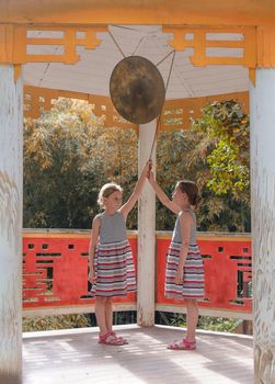 Two tween caucasian girls beating gong in classical asian pavilion near autumn colored trees