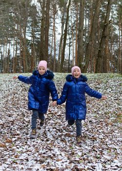 The first frost on autumn. Two happy twin girls in blue coats hand in hand runing down the hill covered with the dry leaves and first snow in the forest