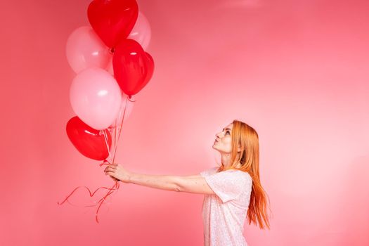 Beautiful redhead girl with red heart baloon posing. Happy Valentine's Day concept. Studio photo of beautiful ginger girl dancing on pink background.