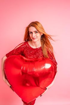 Beautiful redhead girl with red heart baloon posing. Happy Valentine's Day concept. Studio photo of beautiful ginger girl dancing on pink background.