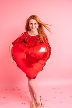 Beautiful redhead girl with red heart baloon posing. Happy Valentine's Day concept. Studio photo of beautiful ginger girl dancing on pink background.