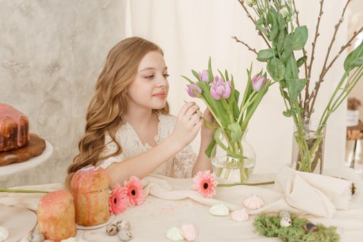 A girl with long hair in a light dress is sitting at the Easter table with cakes, spring flowers and quail eggs. Happy Easter celebration