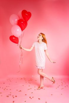 Beautiful redhead girl with red heart baloon posing. Happy Valentine's Day concept. Studio photo of beautiful ginger girl dancing on pink background.