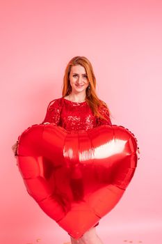 Beautiful redhead girl with red heart baloon posing. Happy Valentine's Day concept. Studio photo of beautiful ginger girl dancing on pink background.