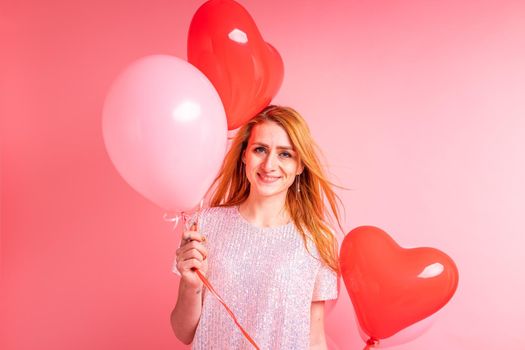 Beautiful redhead girl with red heart baloon posing. Happy Valentine's Day concept. Studio photo of beautiful ginger girl dancing on pink background.