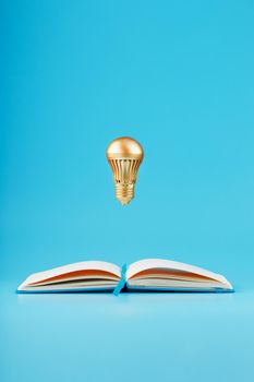 A gold-colored light bulb hangs above the blank pages of a notebook on a blue background. Concept of the idea. Minimalism, isolate.