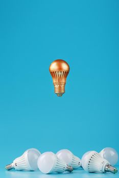 A prominent gold light bulb levitates above an environment of white led bulbs. The concept of an unusual idea and the contrast of surrounding rivals. Minimalism.