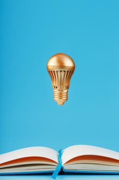 A gold-colored light bulb hangs above the blank pages of a notebook on a blue background. Concept of the idea. Minimalism, isolate.