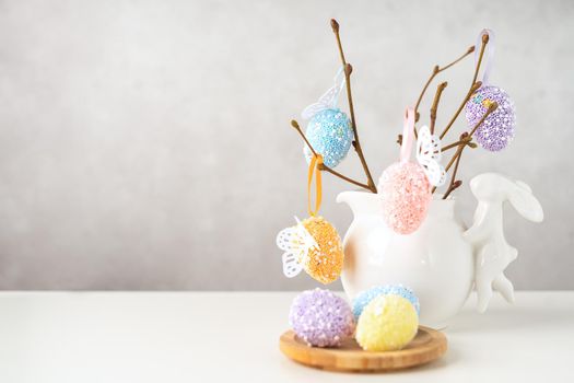 Home interior with easter decor. Vase with willow tree branches with Easter eggs and bunny on white table and background with copy space.