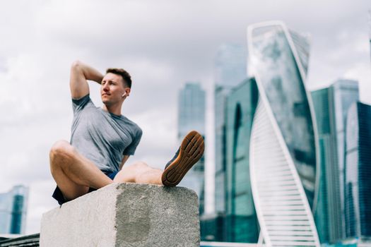 Tired Young man runner sitting on stairs and relaxing after sport training. Holding water bottle while doing fitness workout in summer city urban street, cloudy sky
