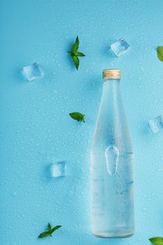 A bottle of ice water, ice cubes, drops and mint leaves on a blue background. Closed glass flask with a drink, layout. Free space, The view from the top