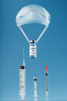 Ampoules with a vaccine against the coronavirus infection COVID-19 were dropped by parachute from a protective mask on a blue background. The concept of assistance in the mass vaccination of the population from the virus. Steamer flight, fast air delivery.